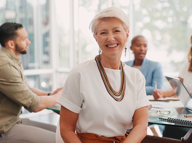 Woman in white blouse with colored necklace smiling at a table with coworkers